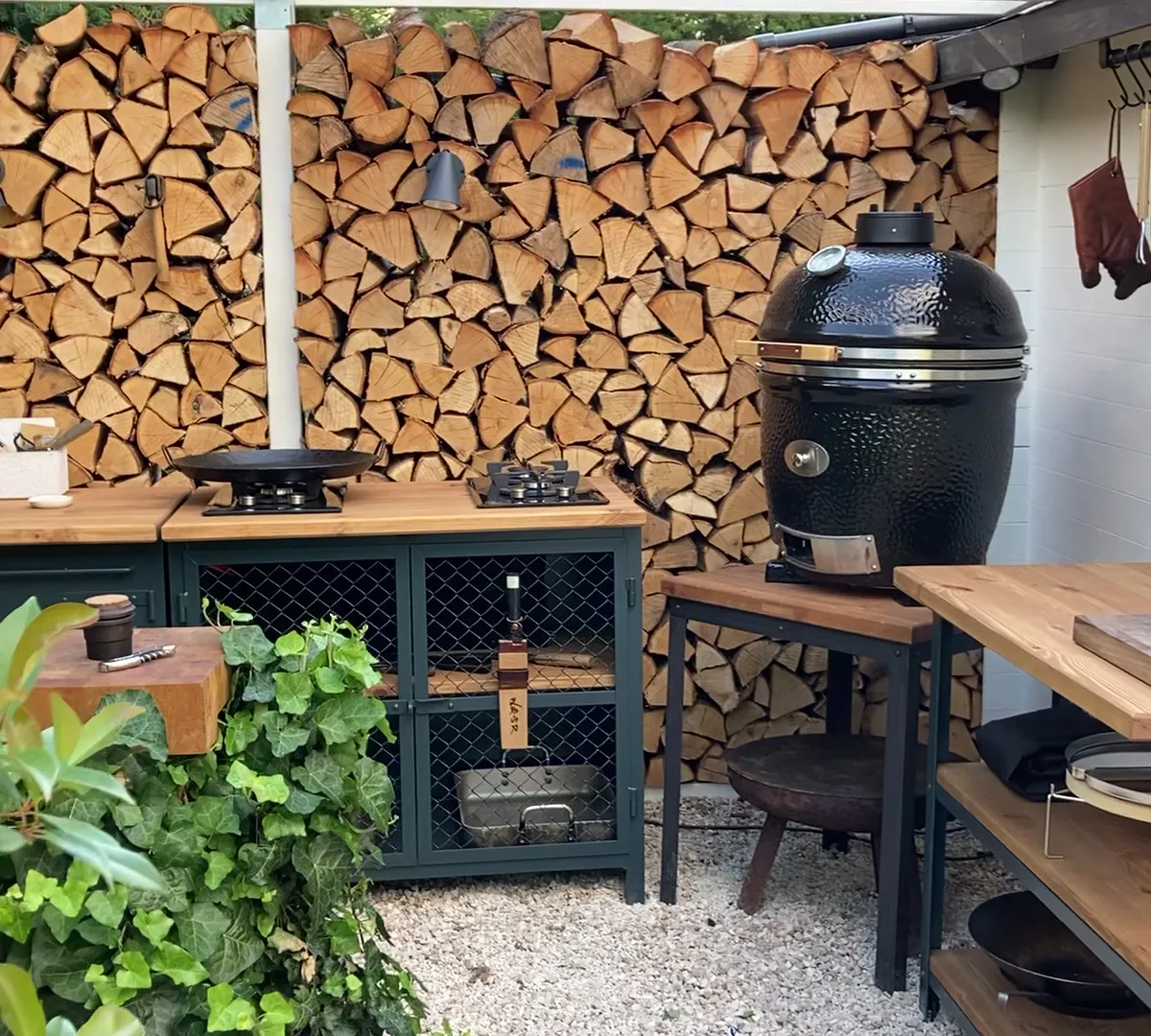 Front view of a bespoke outdoor kitchen by ROUGHKITCHEN Berlin – firewood wall, kamado grill, workbenches and tool setup